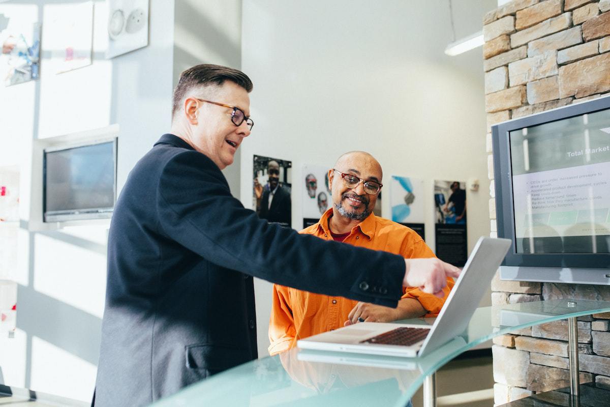 Diálogo: Vendas simples x vendas complexas Dois homens conversando diante de balcão de vidro em interior de prédio analisando algo em notebook sobre este balcão enquanto sorriem e um deles aponta para a tela do aparelho