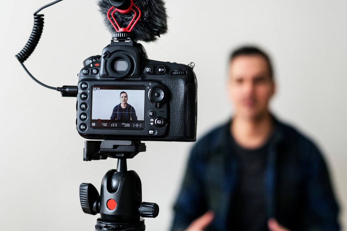 Gravação: Videoaulas para monetizar no YouTube Homem de preto gravando vídeo diante de câmera semi profissional em pedestal com foco na pequena tela do aparelho