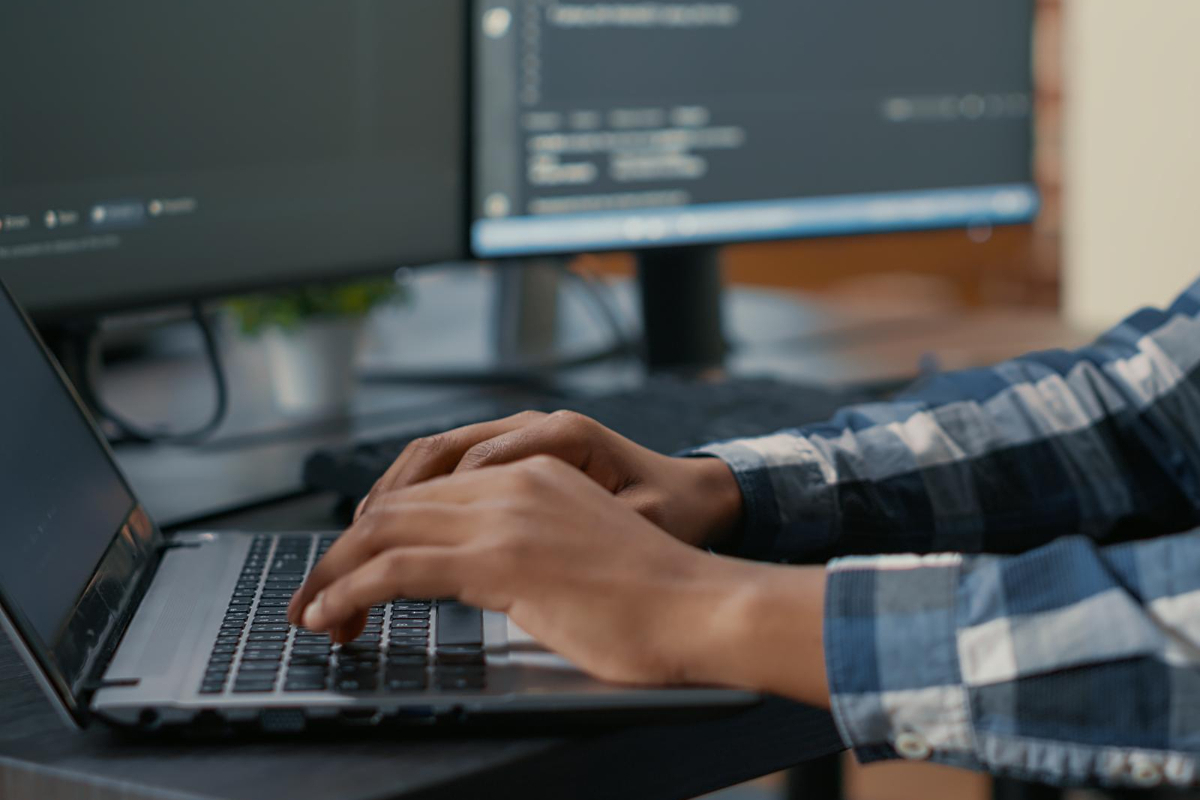 Programador: Web developer Pessoa programando em notebook usando camisa de manga longa xadrez azul com dois grandes monitores à sua direita em desfoco ao fundo