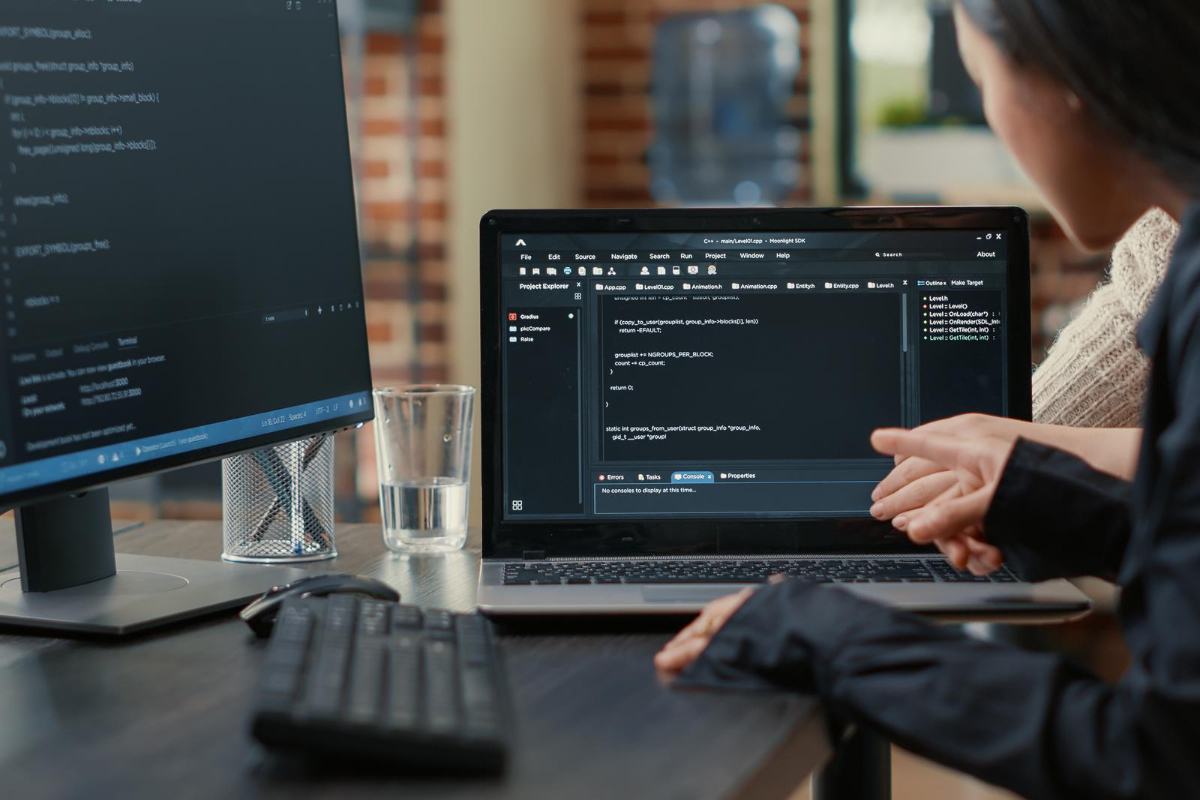 Mulher programando: Web developer Mulher programando em computador preto em cima de mesa de madeira preta enquanto aponta para notebook à sua direita mostrando algo a companheiro de trabalho