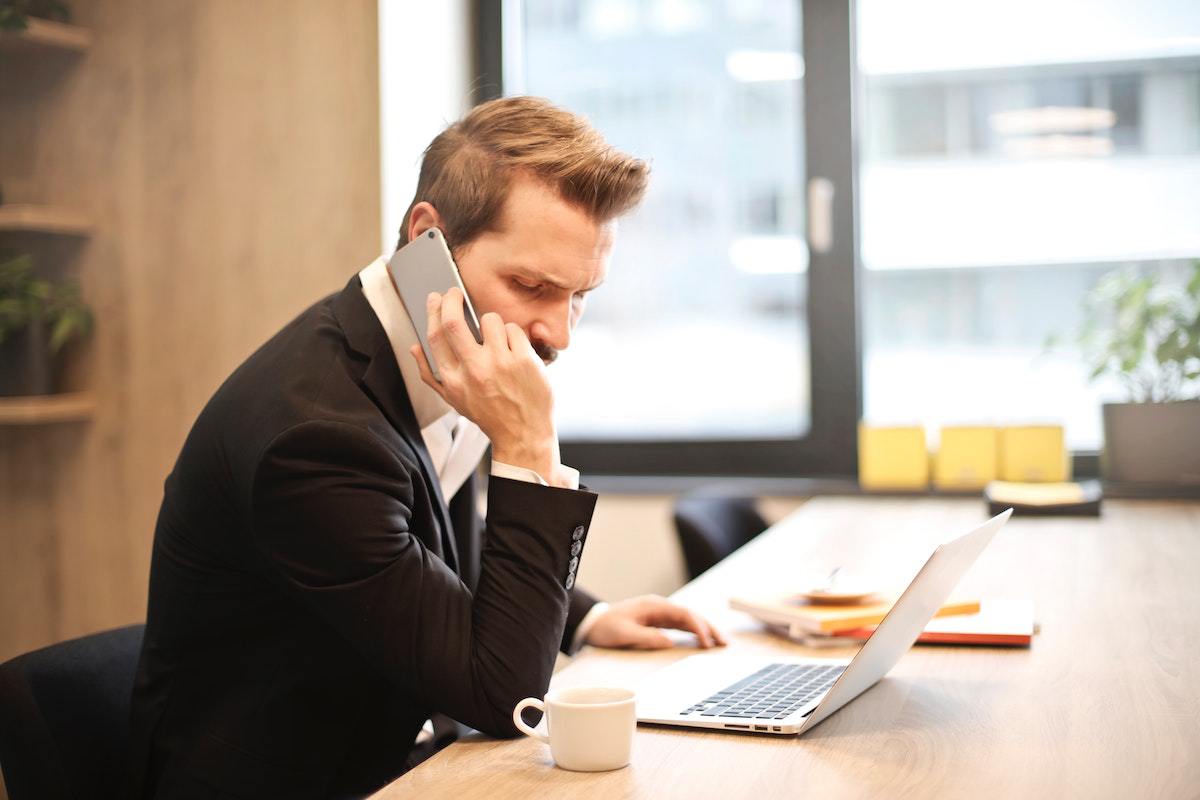 homem sentado, falando ao telefone enquanto trabalha em seu laptop