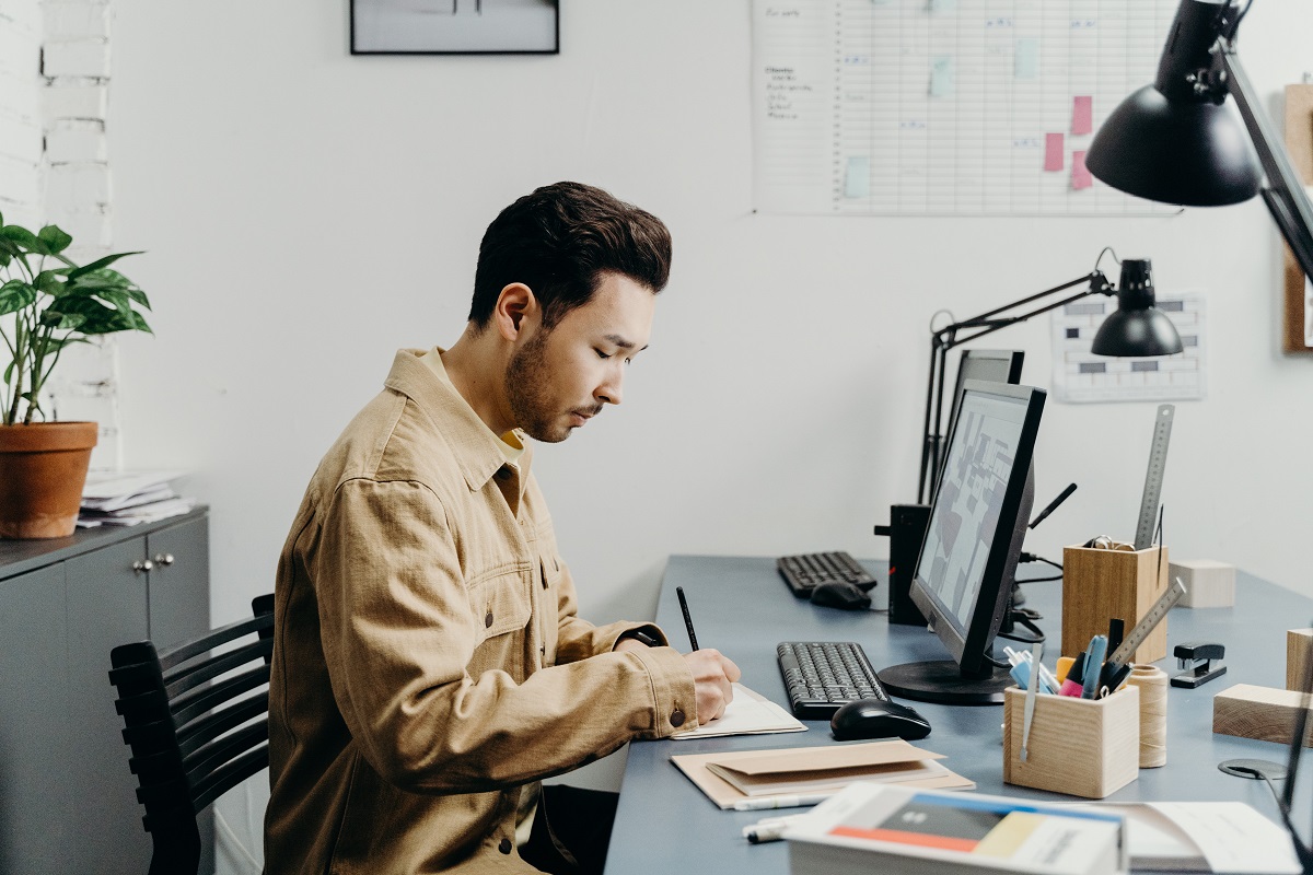 Redação publicitária: A imagem mostra um home na frente de um computador enquanto escreve algo em uma agenda