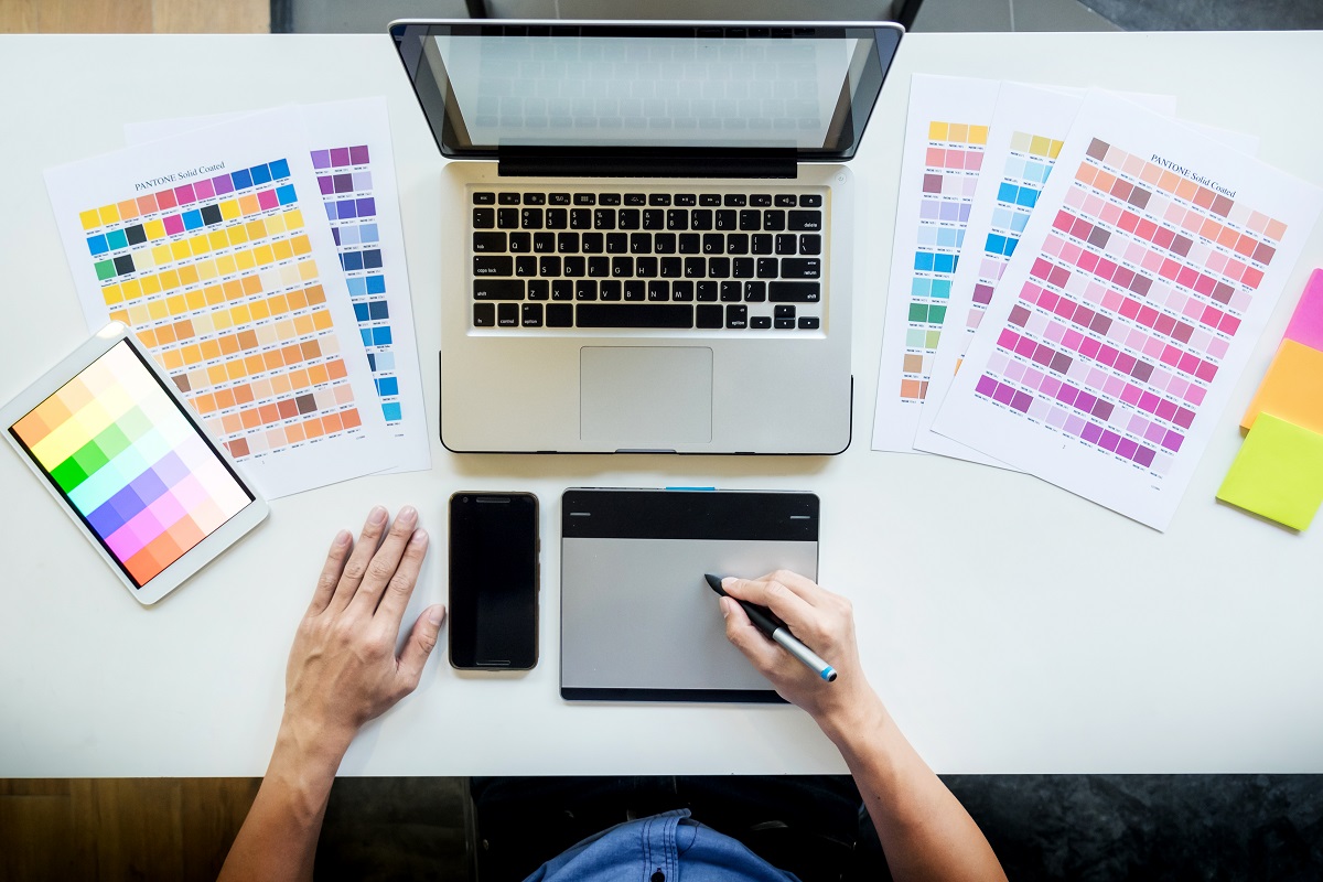 Tendências de design perigosas: A imagem mostra um designer fazendo o seu trabalho. Com ele está um computadores e ao redor algumas paletas de cores. 