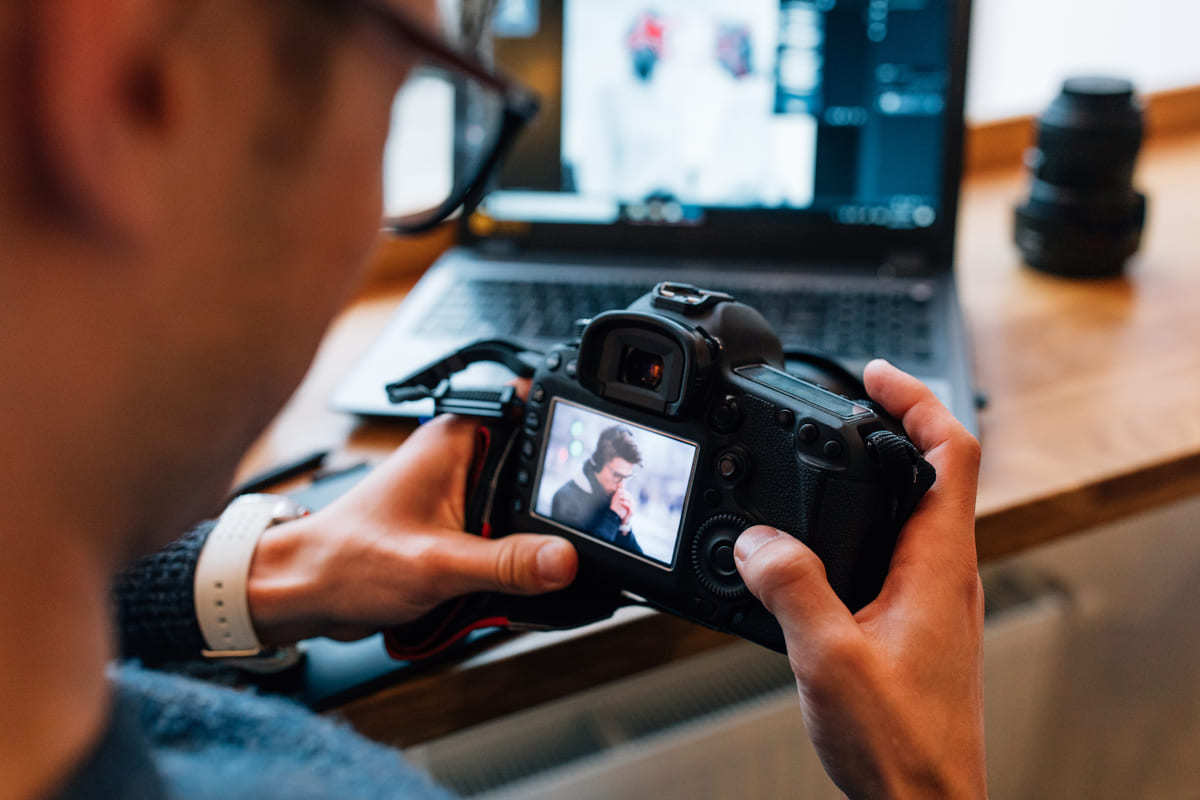 Homem analisando fotografia em tela de câmera fotográfica com notebook ao fundo em desfoco usando Adobe Lightroom sobre mesa de madeira