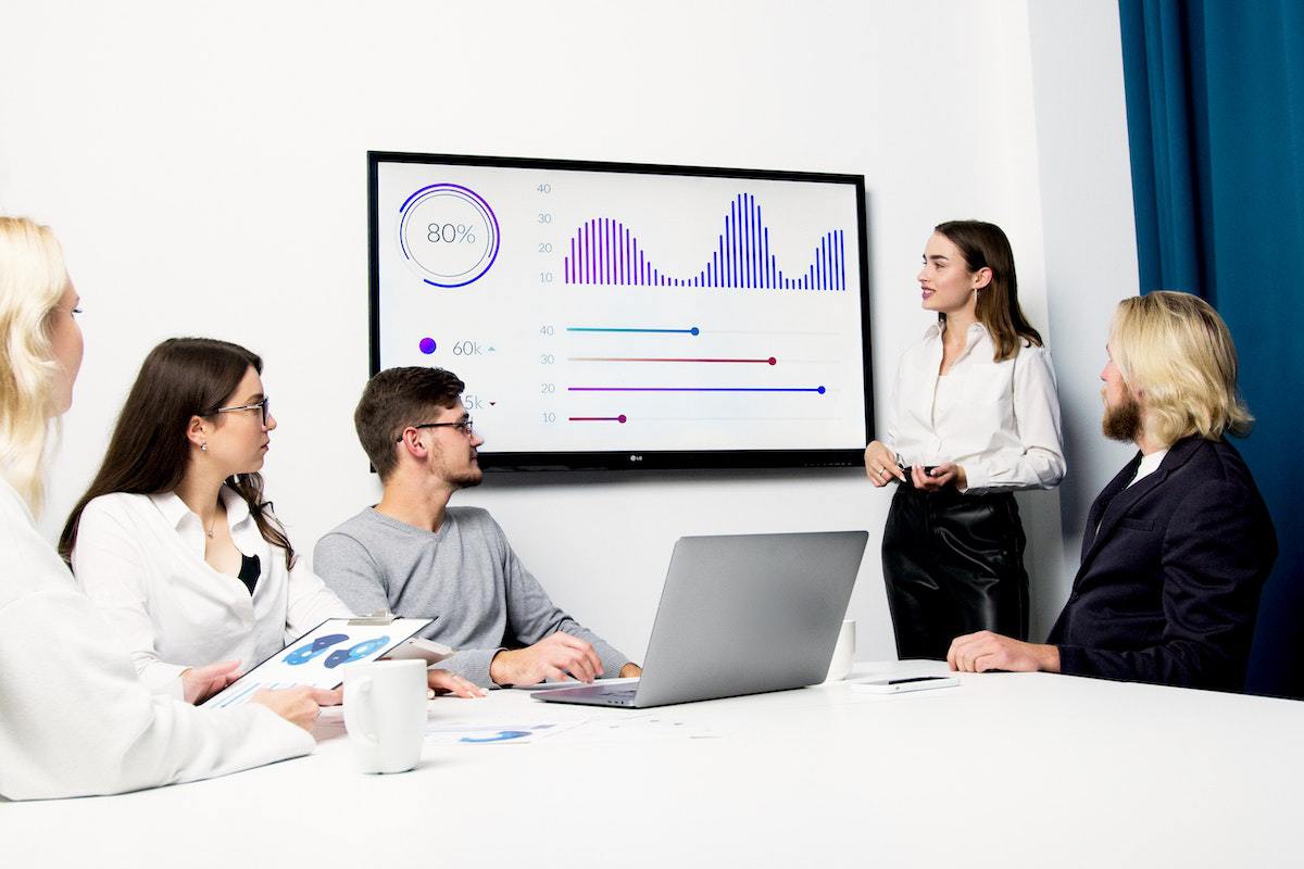 Mulher usando monitor em sala de reuniões para fazer Apresentação no powerpoint para seus colegas com dados e gráficos, onde um deles usa notebook em mesa branca diante da líder