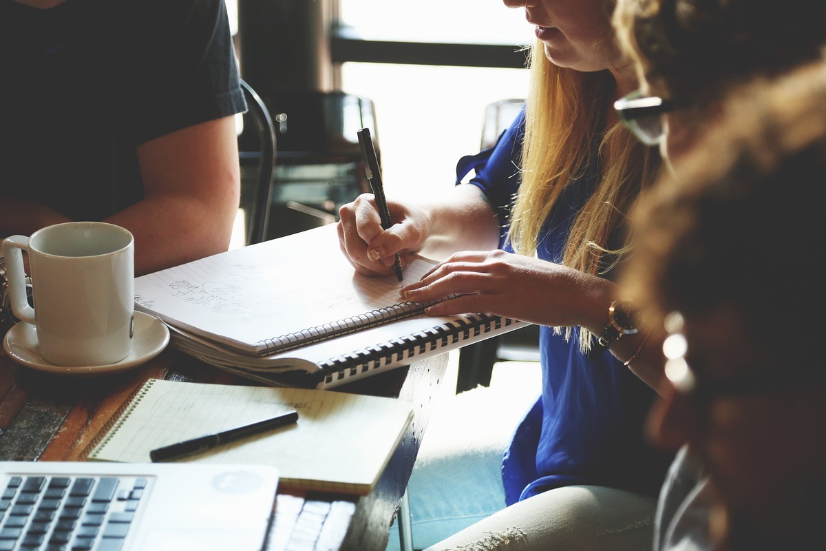Anotações: BV Mulher fazendo anotações em reuniões sobre BV com colegas ao redor de mesa de madeira com notebook e xícara grande sobre a mesma