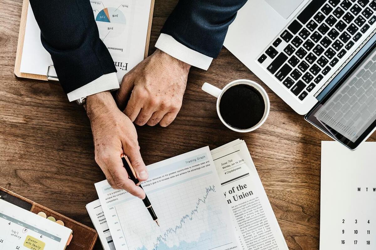 Gráficos: BV Pessoa usando terno diante de mesa de madeira apontando com caneta para gráficos impressos em folha a4 sobre jornal dobrado com caneca de café ao lado do mesmo, além de notebook e mais papéis em pranchetas