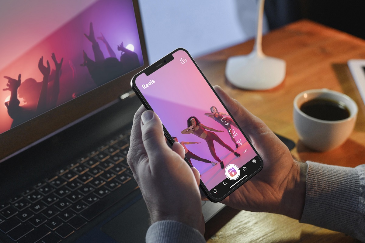 Homem usando smartphone diante de notebook vendo mídia de Comarketing com vídeo com xícara de café ao lado sobre mesa de madeira