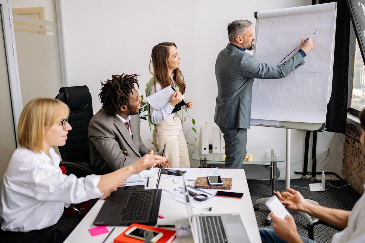 Reunião em local de trabalho com líder anotando algo em painel de cartolinas brancas e com time sentado à sua frente para Criar roteiro matador para webinar