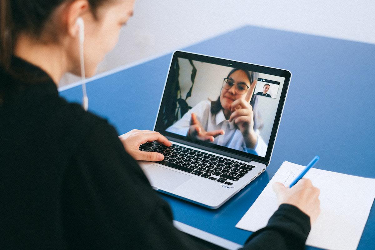 Mulher em reunião online com outra mulher anotando algo em bloco de folhas brancas com notebook sobre mesa azul