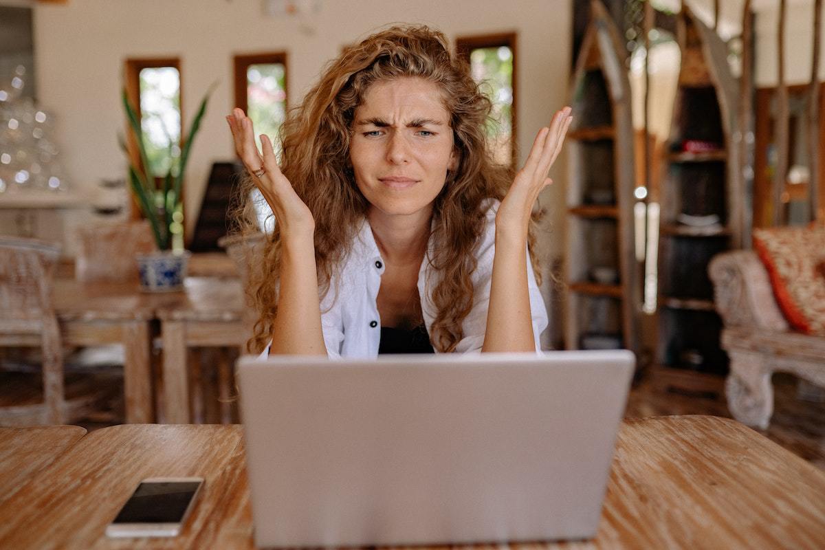 Mulher trabalhando em home office usando notebook sobre mesa de madeira com celular com tela bloqueada à sua direita com ar de confusa e mãos em gesto com as mãos como se estivesse se perguntando algo