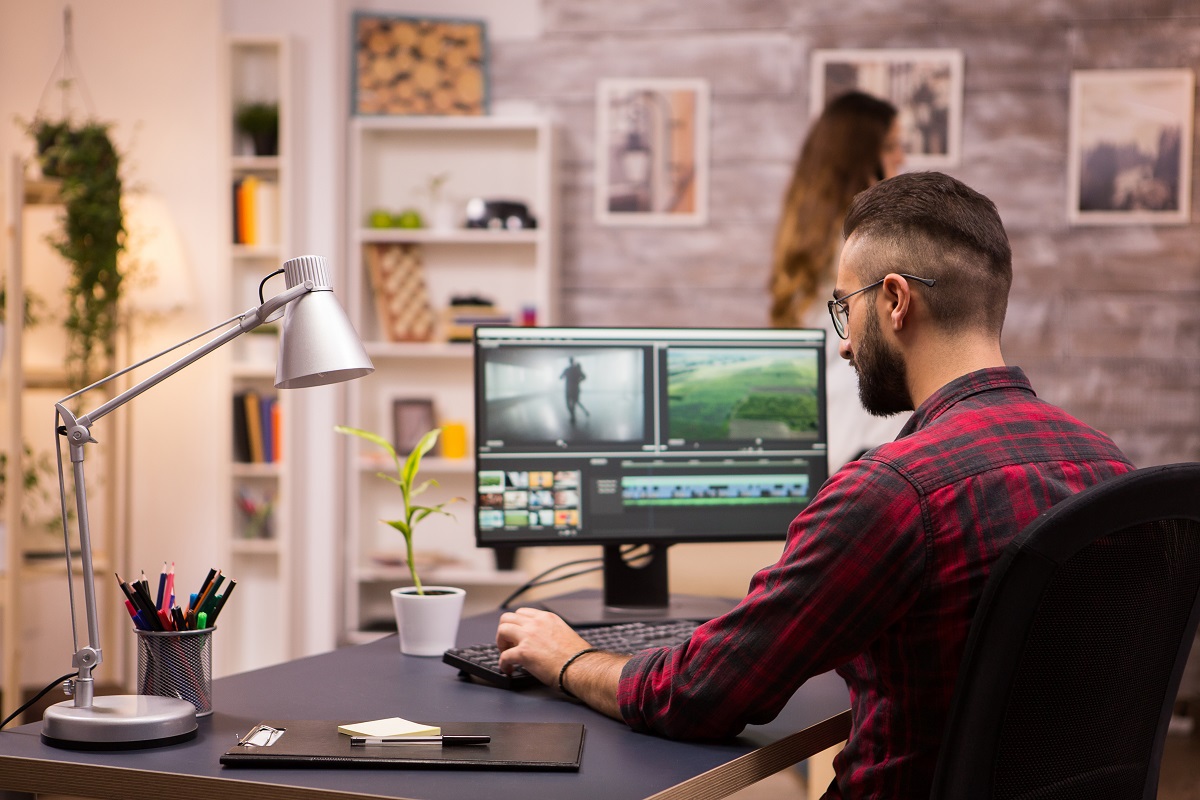 Homem trabalhando em mesa preta editando imagens em seu computador com abajur ao lado e mulher ao fundo