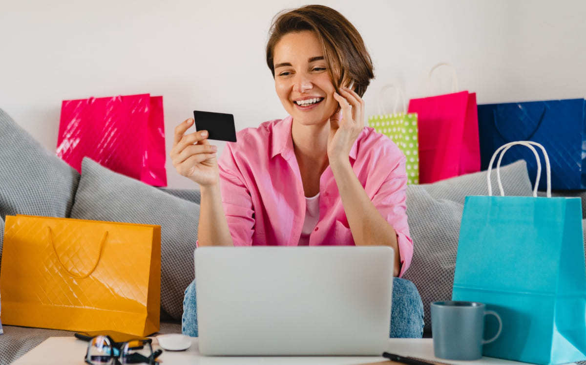Cartão: E-commerce de serviços B2B Mulher sentada em sofá diante de mesa de centro com notebook cinza em cima do mesmo sorridente e olhando para cartão de créditos com sacolas espalhadas ao redor
