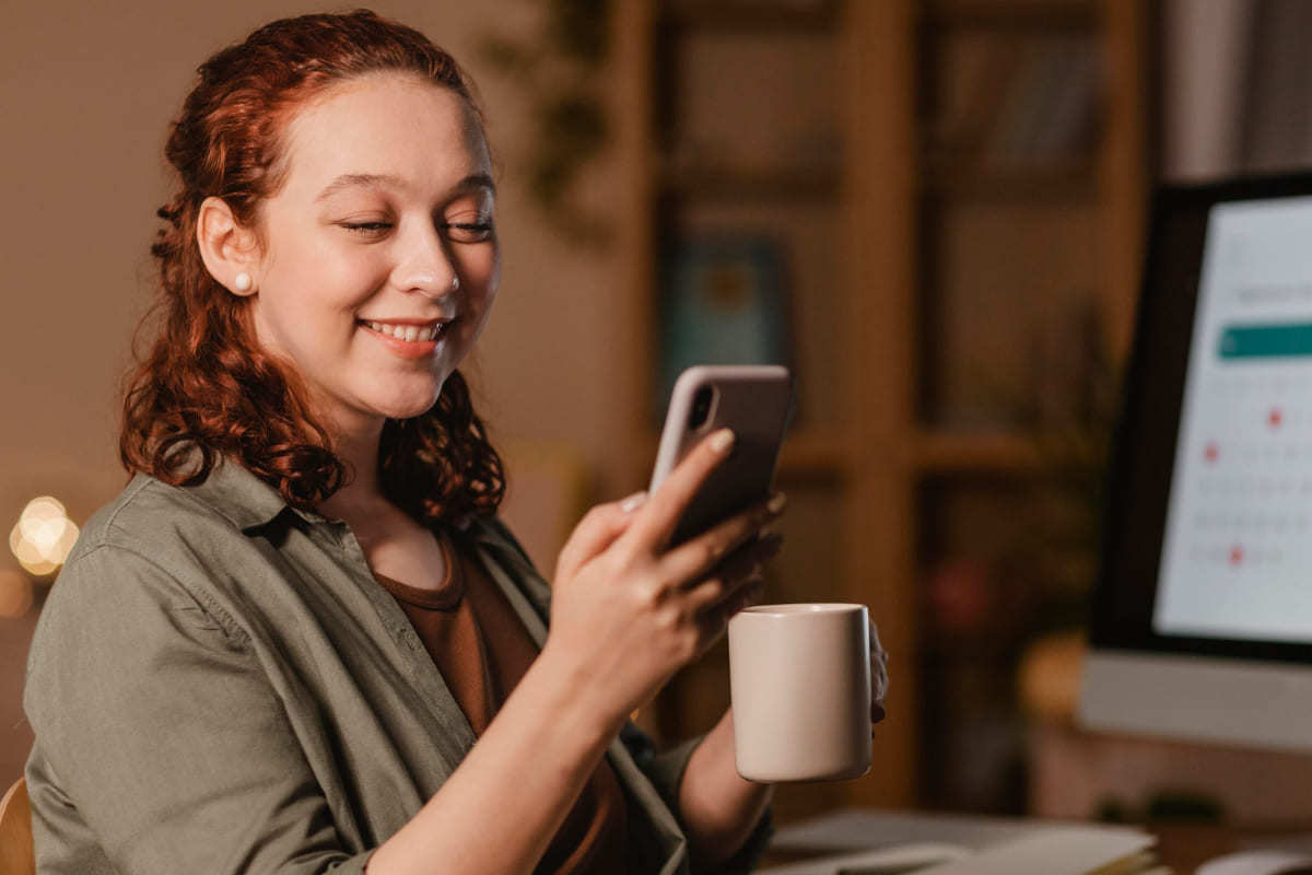 Aparelhos: E-commerce de serviços B2B Mulher sorridente usando celular sentada de frente à mesa com computador e analisando aparelhos com E-commerce de serviços B2B