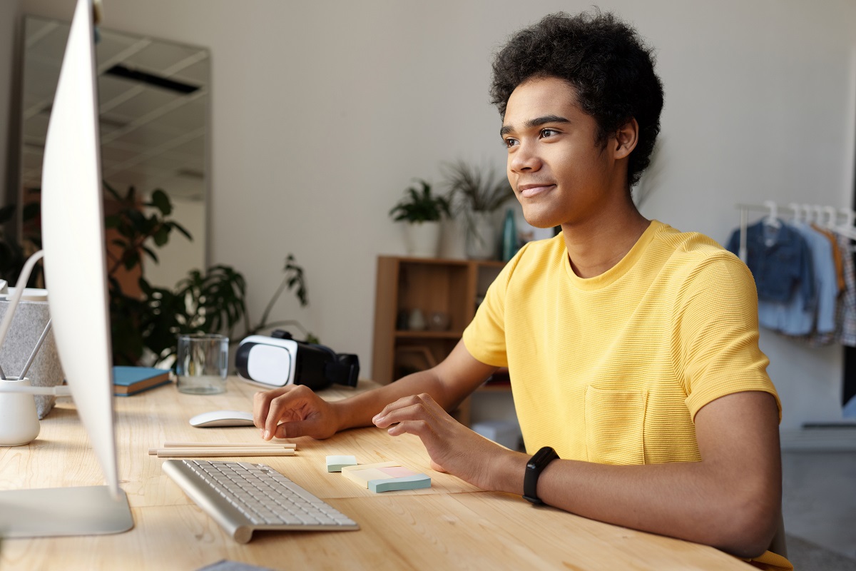 Homem jovem sentado diante de computador sobre mesa de madeira assistindo aula na modalidade EAD