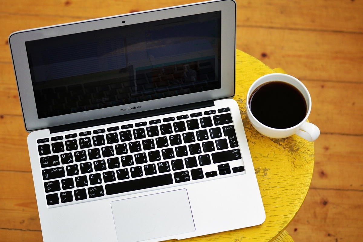 Notebook Apple modelo Macbook Air sobre mesa pequena redonda de madeira ao lado de xícara de café e ao lado de xícara de café branca bem cheia e com piso também de madeira ao fundo da foto, tirada em ângulo de cima para baixo