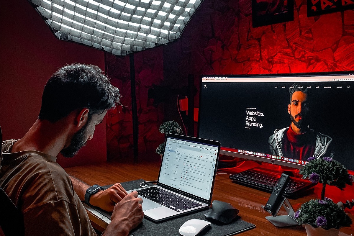 Homem trabalhando com notebook analisando seu e-mail com monitor grande ao fundo com montagem com sua foto em ambiente com iluminação vermelha
