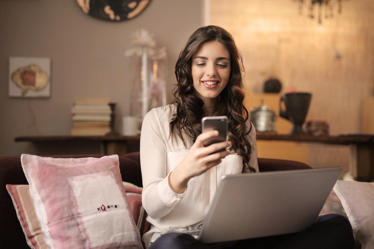 Mulher sentada diante de notebook usando celular como Estratégias de vendas ao lado de almofadas em sala de sua casa