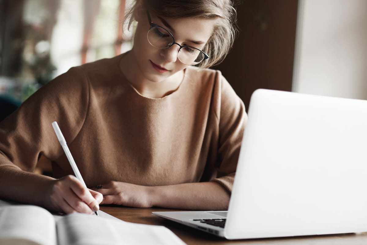 Mulher trabalhando em notebook sobre mesa de madeira anotando algo em agenda e usando Extensões Google Chrome em home office
