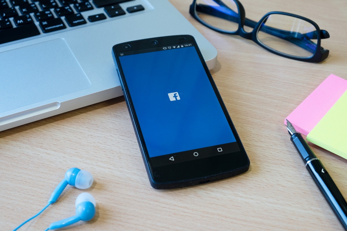 Aplicativo do Facebook em tela inicial em celular apoiado em teclado de notebook sobre mesa de madeira e ao lado de outros objetos