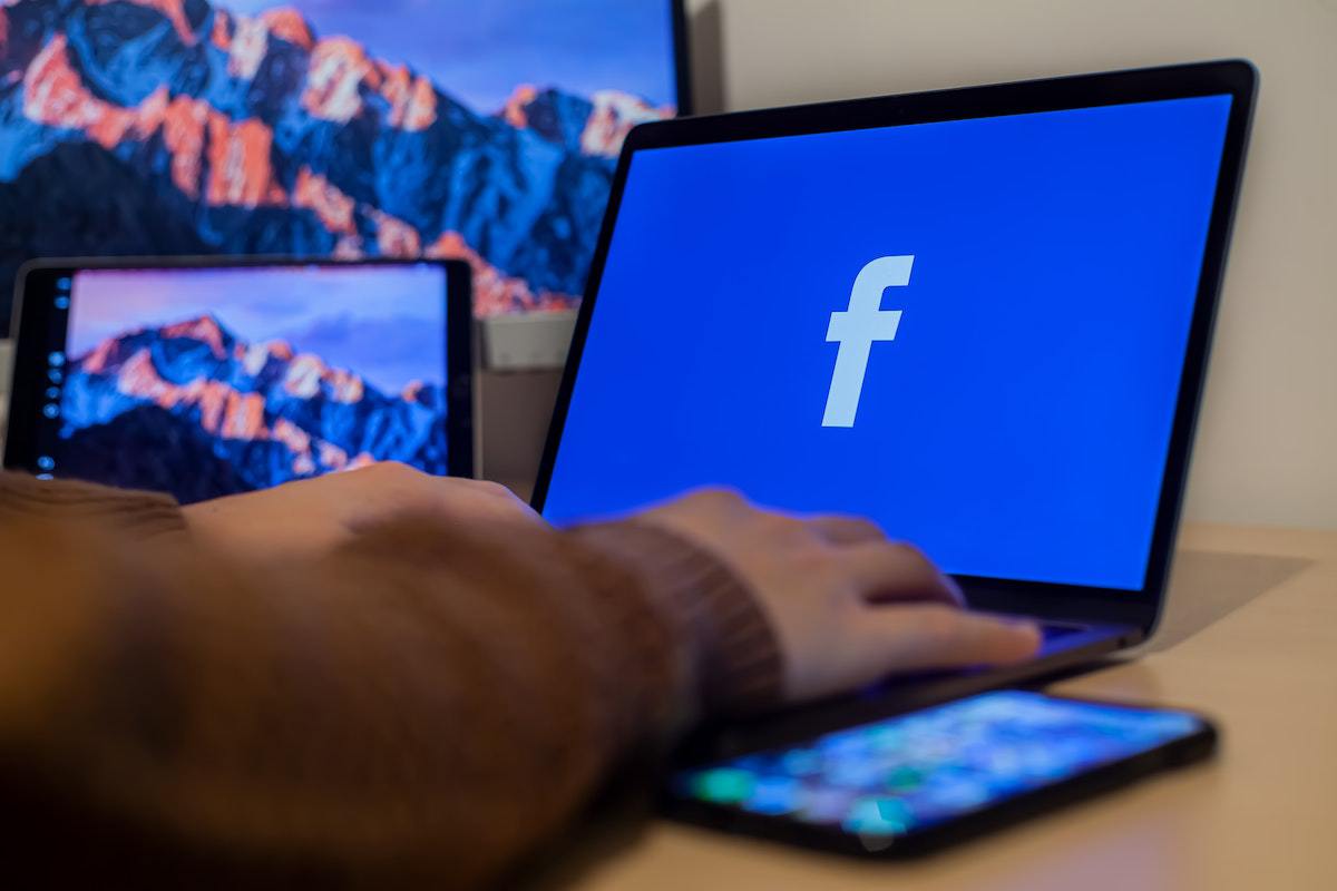 Pessoa acessando Facebook com smartphone do lado, além de tablet com monitor ao fundo da imagem em desfoco ao fundo