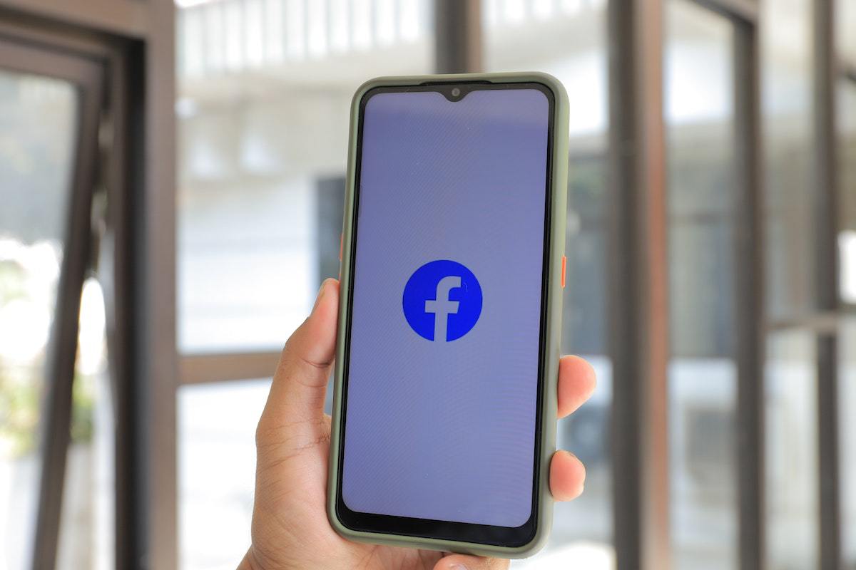 Pessoa segurando smartphone com tela de processamento de abertura do Facebook diante de janela de vidro de edifício