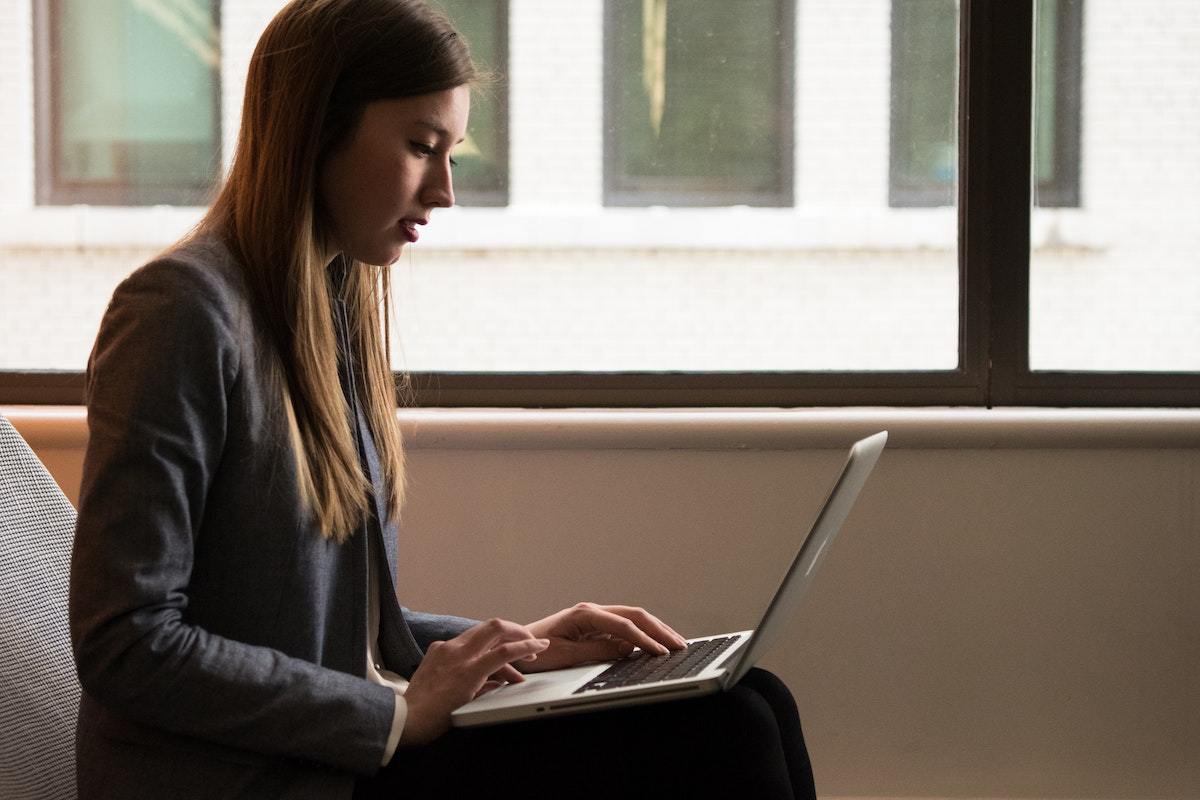 Mulher sentada com notebook no colo diante de janela de vidro de prédio