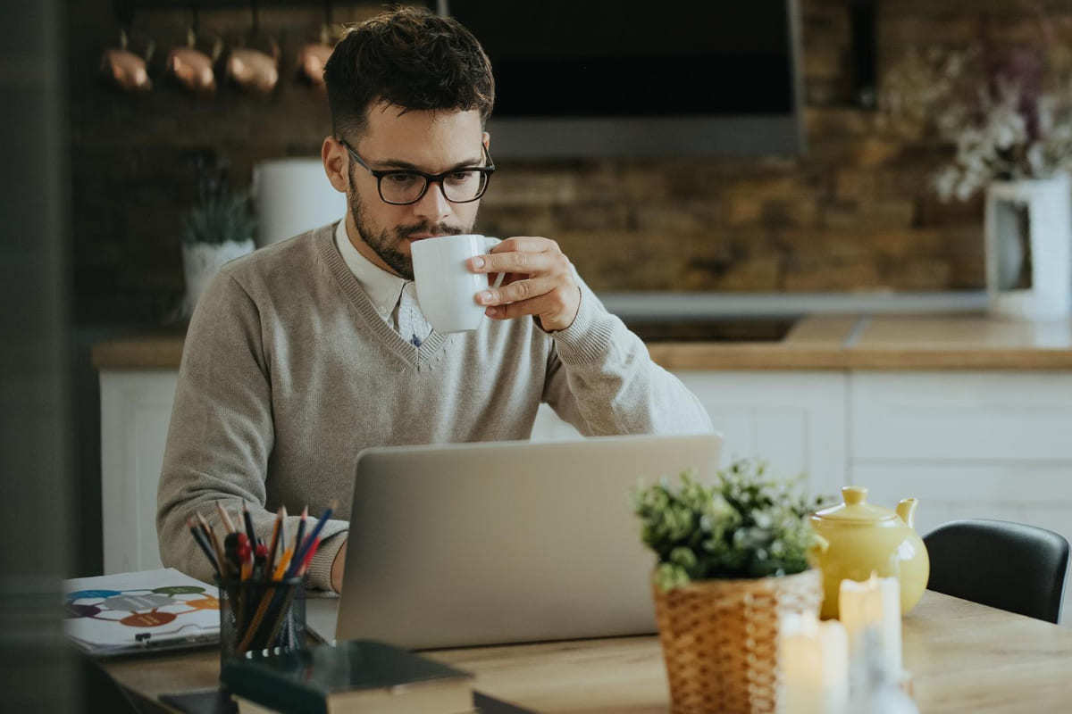 Home office: Gerenciar canal do YouTube Homem trabalhando em home office com notebook sobre mesa de madeira com planta de interior por trás do aparelho e porta canetas à sua direita enquanto toma bebida em caneca branca