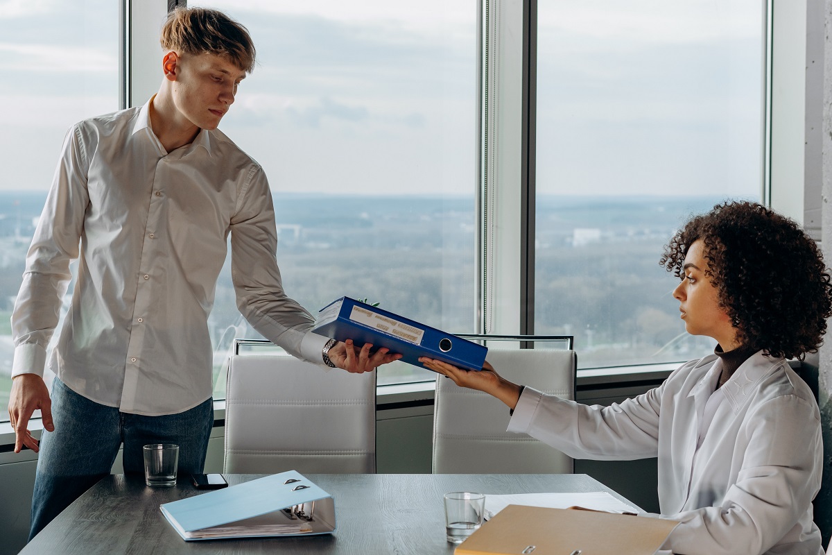 Local de trabalho: Gestão integrada Mulher de jaleco sentada diante de mesa em local de trabalho entregando pasta azul à rapaz de pé diante de janelas de vidro em edifício em empresa que trabalha com Gestão integrada