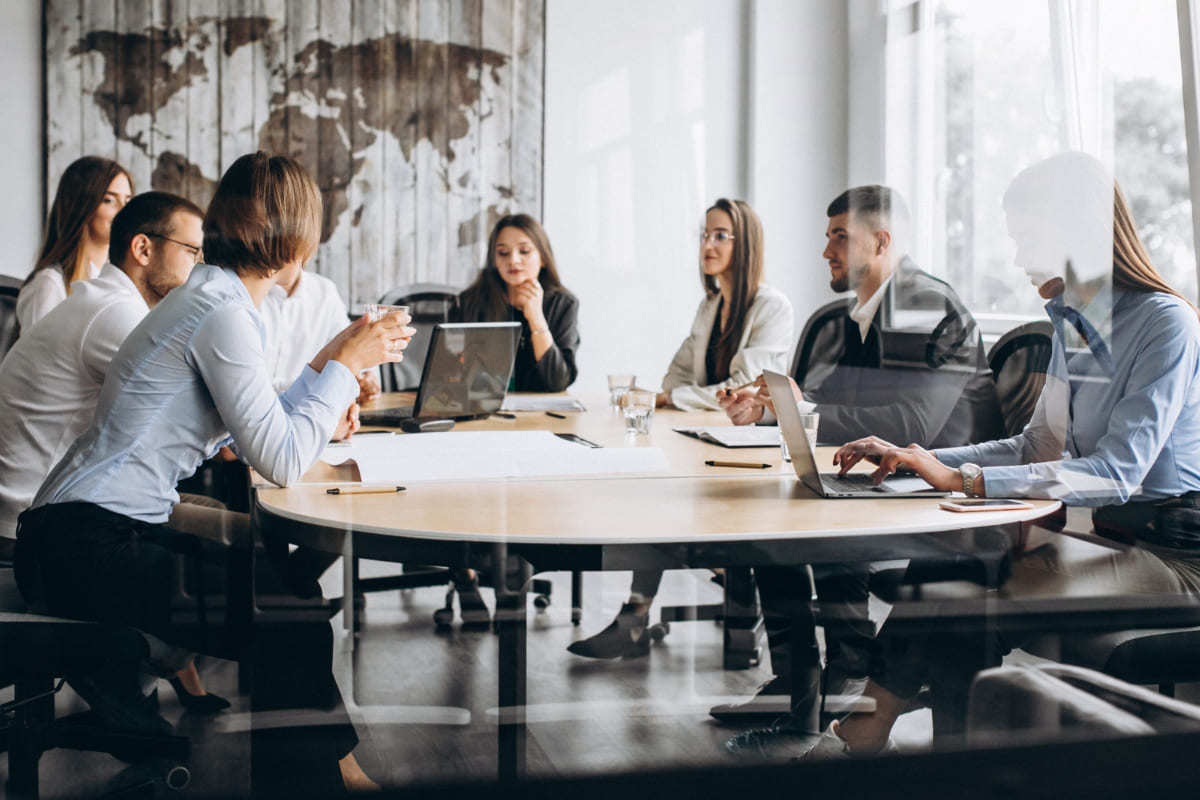 Reunião em local de trabalho com equipe de homens e mulheres debatendo algo ao redor de mesa de madeira analisando dados em aparelhos como notebooks