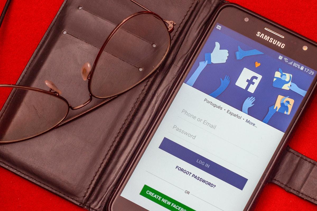 Tela de login do Facebook em smartphone dentro de case de material análogo à couro aberta com cases para cartão de crédito e óculos de grau sobre a mesma