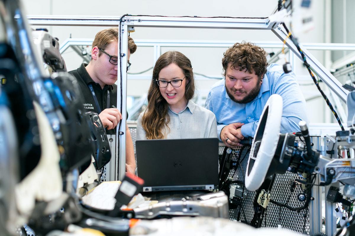Dois homem e uma mulher usando notebook em Indústria 4.0 de automóveis programando algo em interior de carro, parcialmente montado