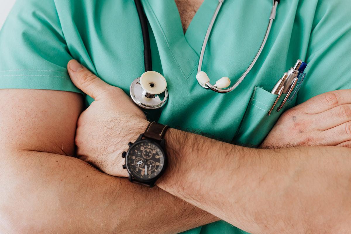 Médico de braços cruzados com estetoscópio no mesmo e relógio preto no pulso esquerdo com roupa verde de ala hospitalar pronto para plantão