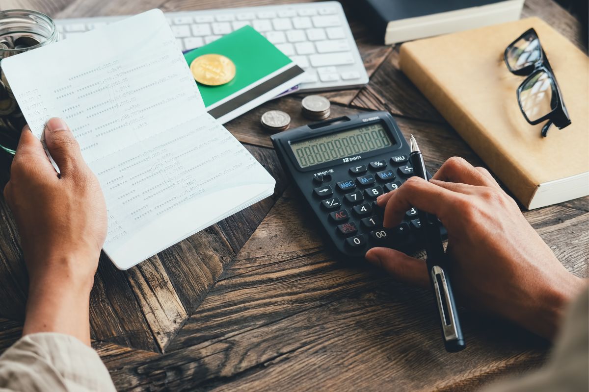 Pessoa sentada diante de mesa de madeira somando lucro em calculadora com folhas na mão e agenda e utensílios de escritório espalhados ao seu redor