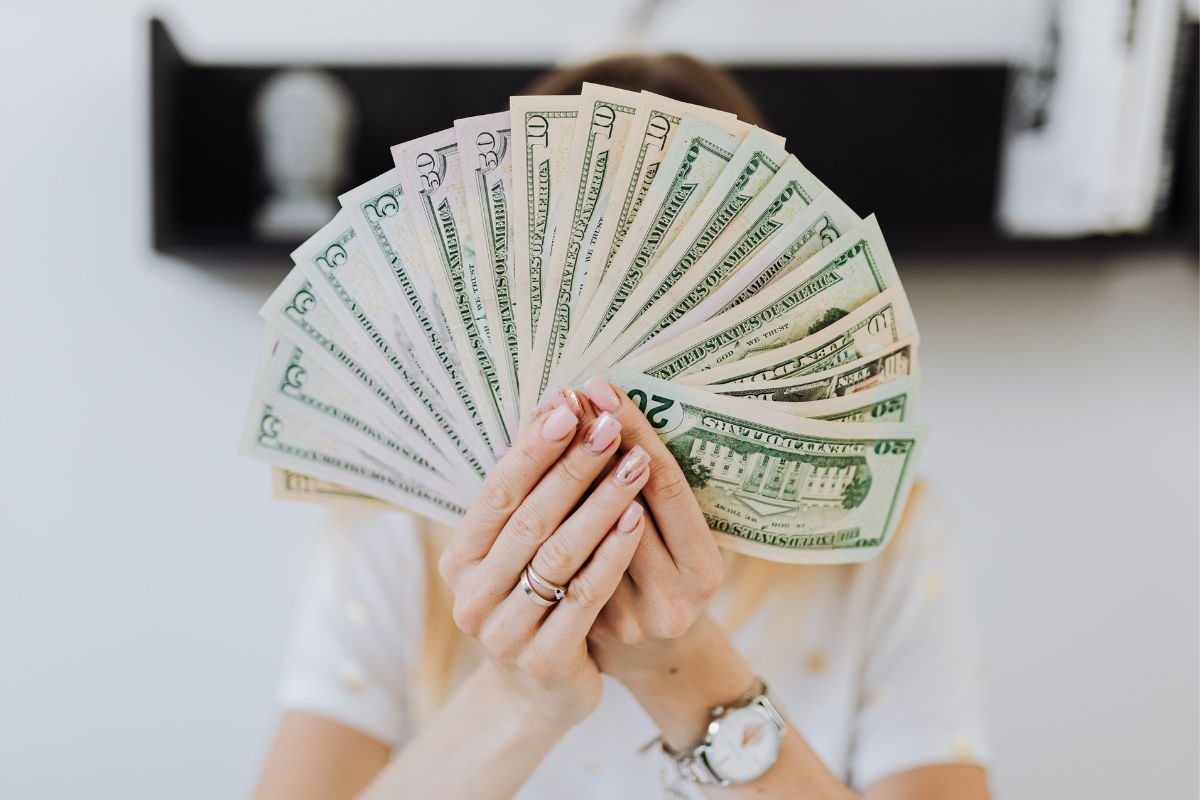 Dinheiro: Lucro Mulher segurando cédulas de dinheiro do seu Lucro diante do seu rosto com relógio prateado no pulso e móvel fixado na parede ao fundo da imagem