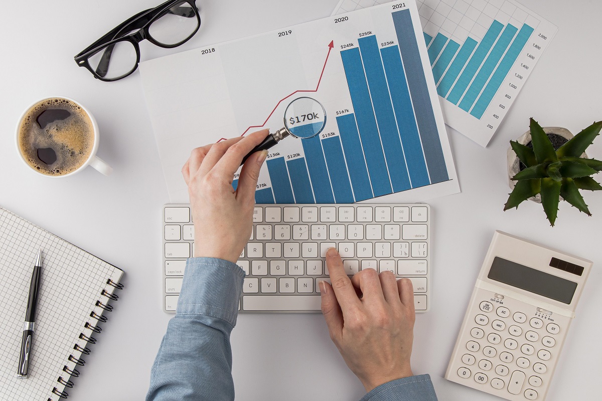 Pessoa em local de trabalho analisando gráficos impressos com lucro em folha com lupa e digitando algo em teclado sem fio branco de computador, com caneca de café à esquerda e óculos de grau dobrado