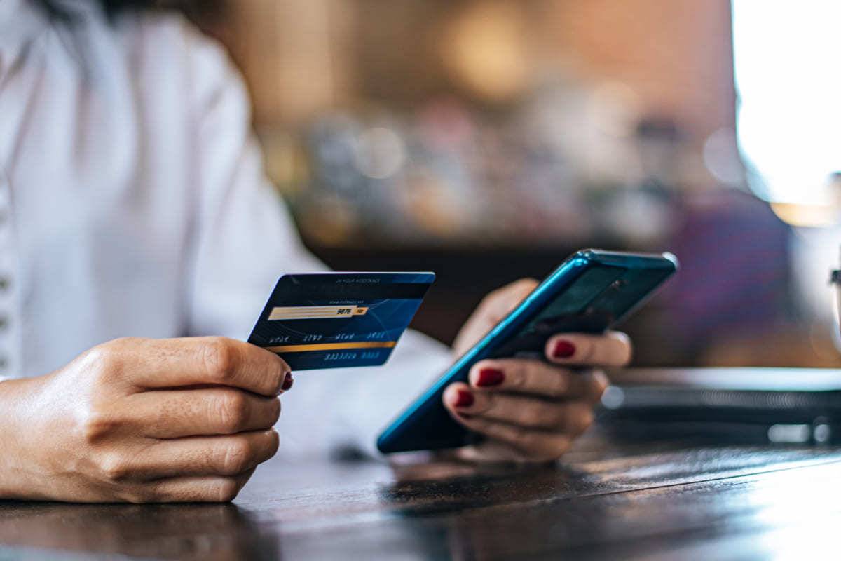 Cartão: Maiores fintechs Pessoa segurando celular com mão esquerda e cartão com mão direita sobre mesa de restaurante