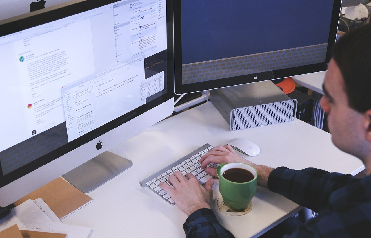 Homem trabalhando em empresa diante de mesa com computador desktop e dois monitores à sua frente