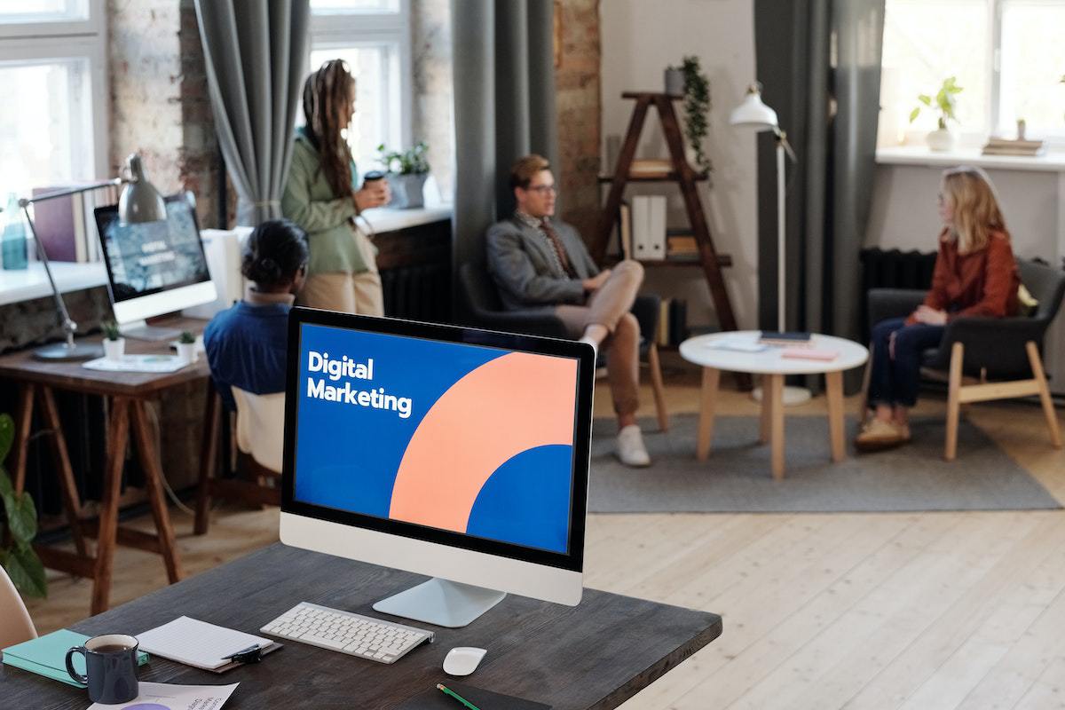 Computador: Matemarketing Computador sobre mesa em local de trabalho com porta lápis na sua frente e caneca preta com montagem na tela escrito Digital Marketing e colegas ao fundo conversando diante de janela de vidro