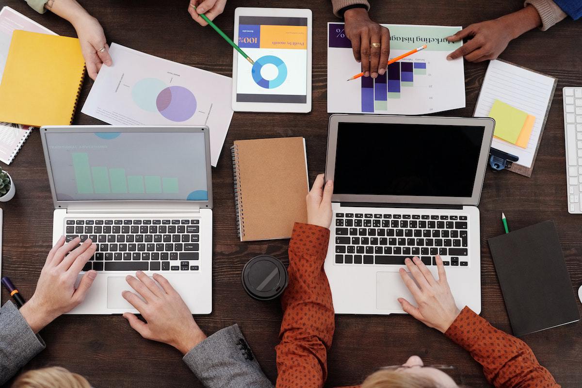 Pessoas trabalhando ao redor de mesa de madeira usando notebook e analisando gráficos em folhas impressas