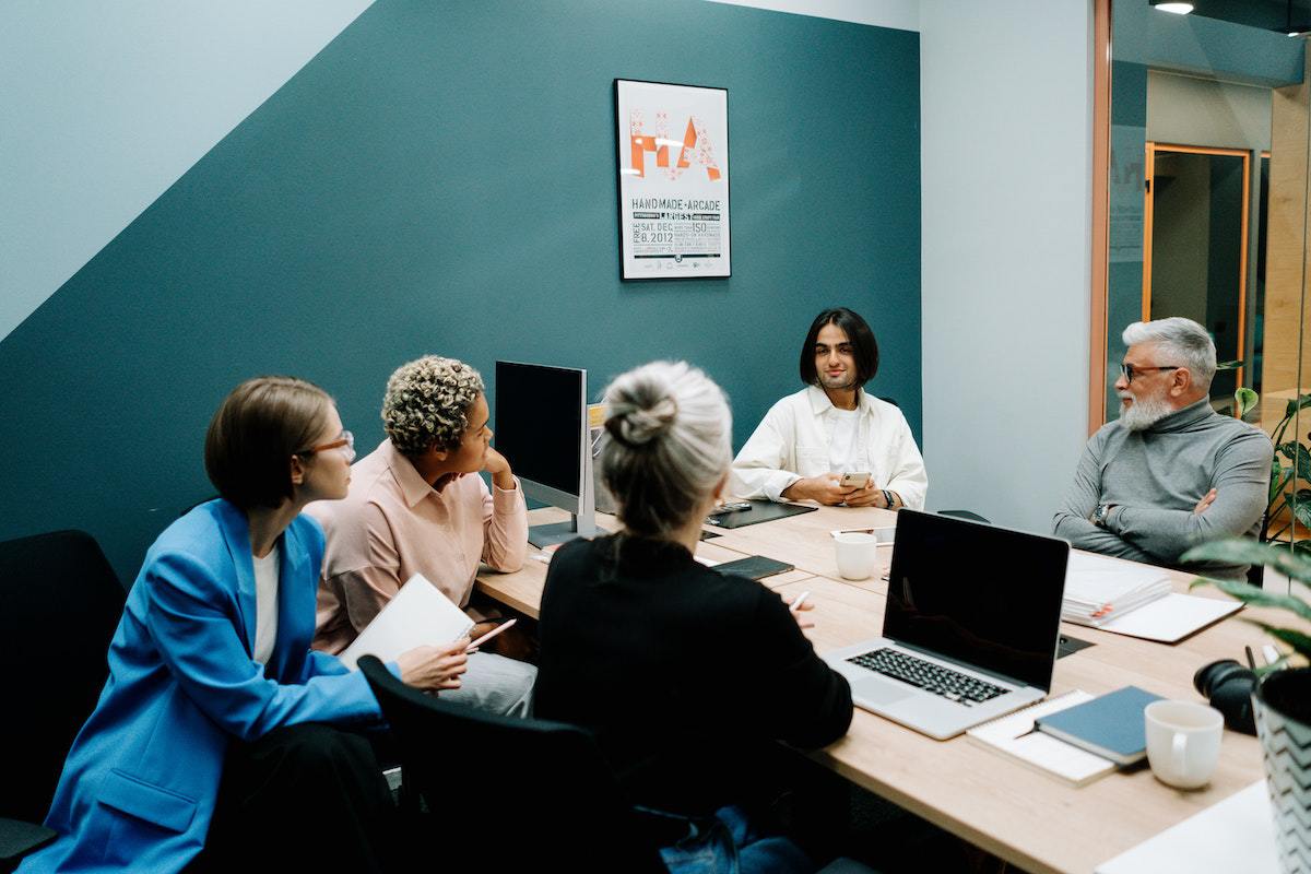 Equipe de trabalho diversa com três mulheres e dois homens sentados ao redor de mesa e usando notebooks e agendas em reunião