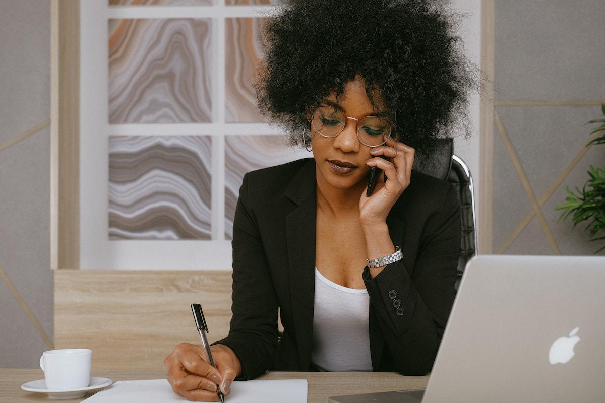 Mulher falando ao telefone sentada diante de mesa enquanto ano algo em papel com notebook à sua frente fazendo negócios
