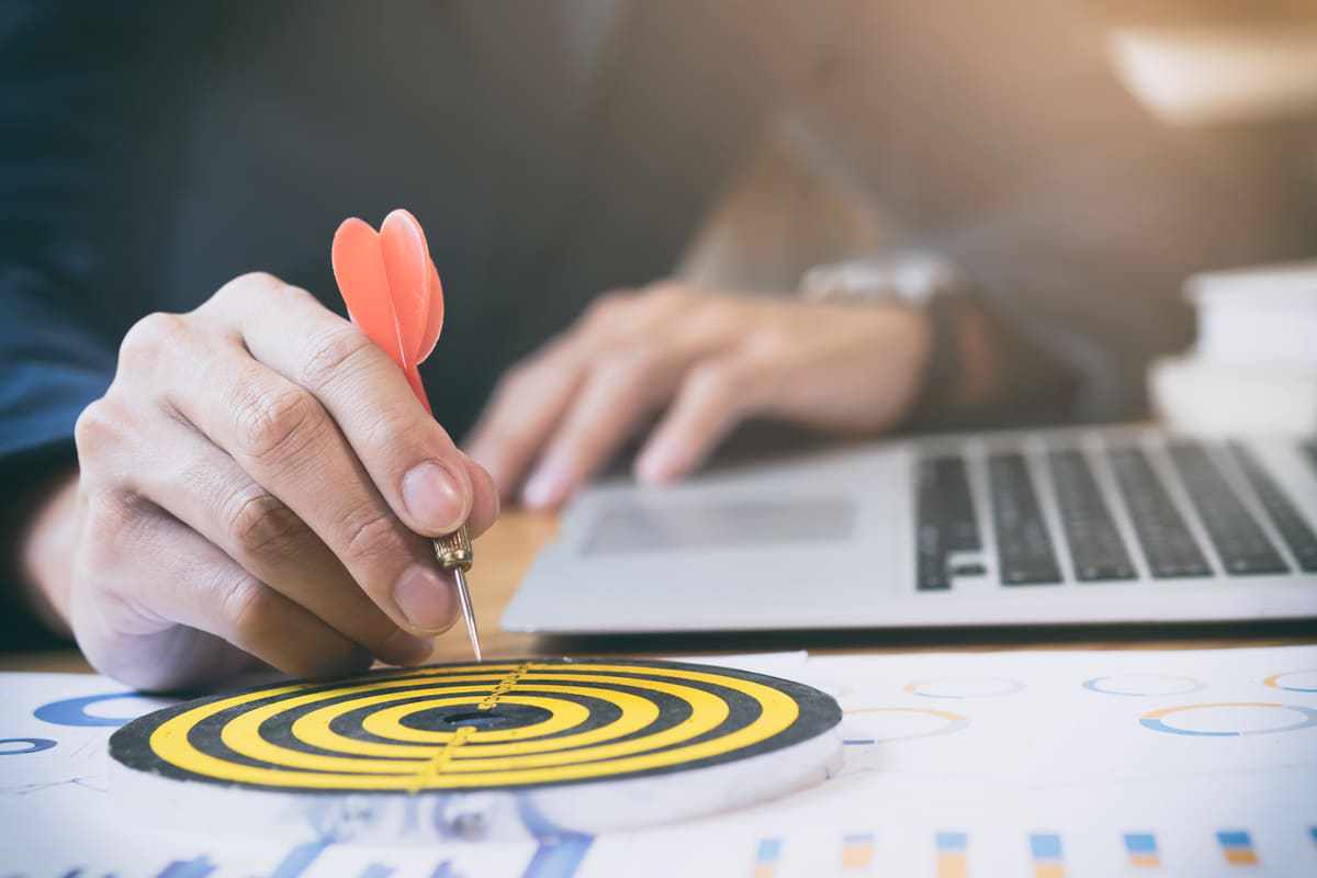 Pessoa usando notebook com alvo ao seu lado direito e dados impressos com dardo enquanto o coloca no centro do alvo impresso