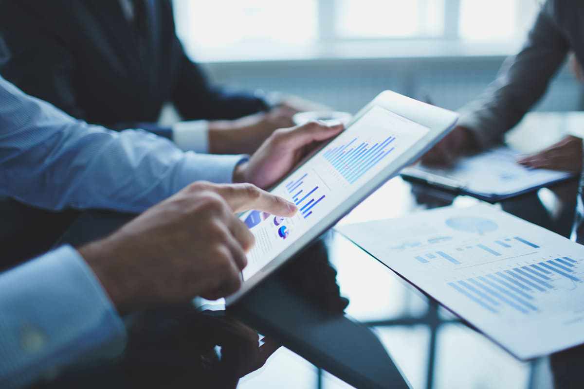 Duas pessoas em escritório analisando dados impressos em folhas sobre mesa de vidro e um deles conferindo gráficos em tablet branco