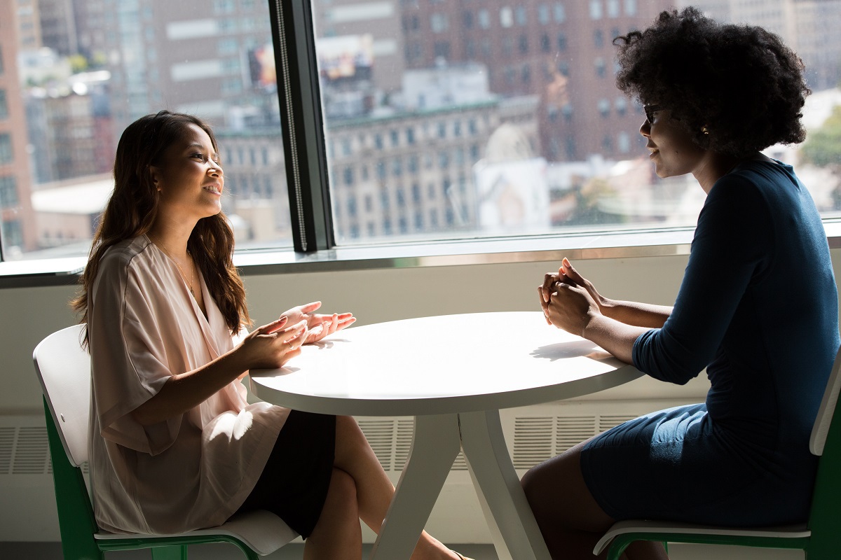 Duas mulheres em reunião sentadas frente a frente diante de mesa de madeira redonda ao lado de janela de vidro de edifício