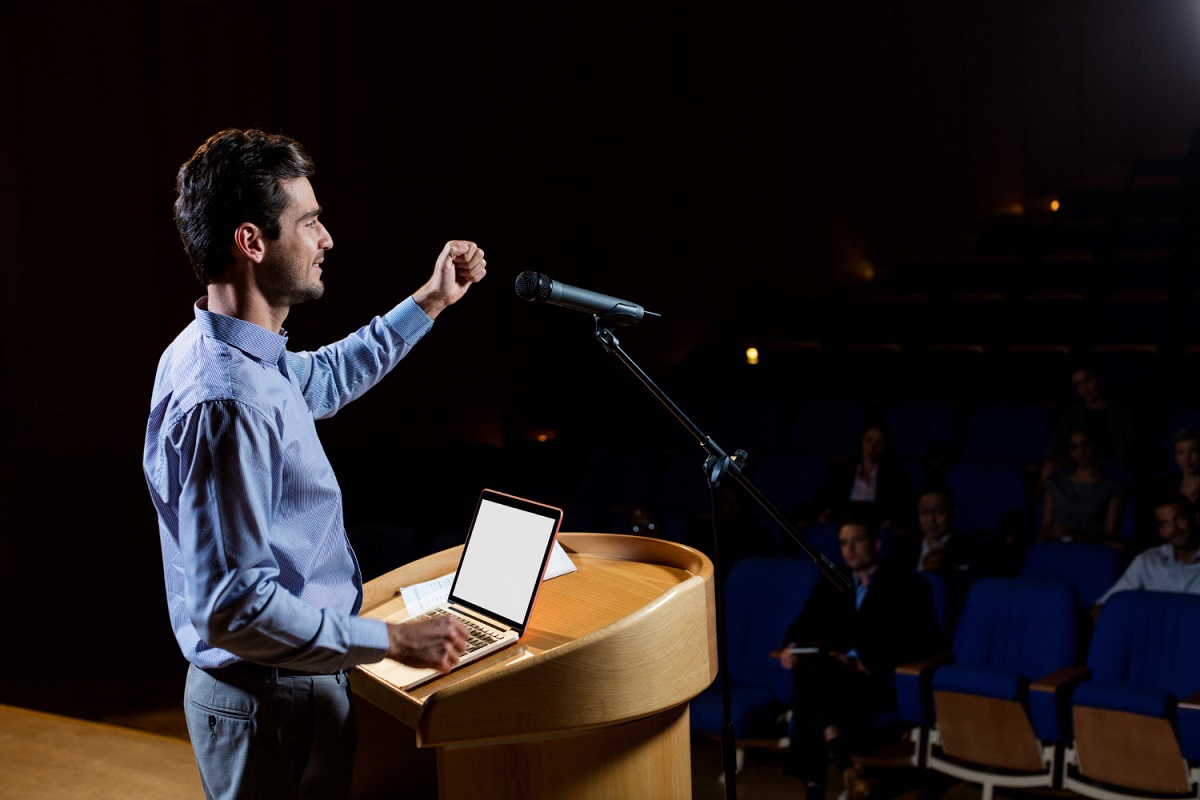 Palestra: Oratória Homem durante palestra em Oratória em local análogo à teatro com notebook sobre palanco falando em microfone para plateia à sua frente e abaixo