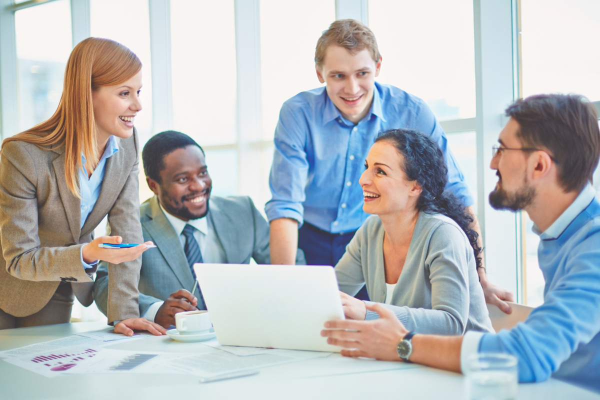 Duas mulheres e três homens sorridentes em sala de reunião analisando dados impressos em folhas a4 e notebook, diante de janela de de edifício