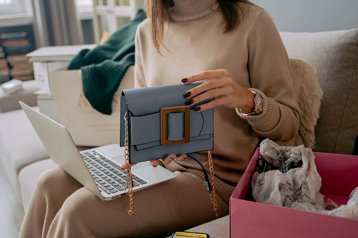 Mulher com notebook no colo e escolher objetos como bolsas enquanto senta em sofá no interior de casa
