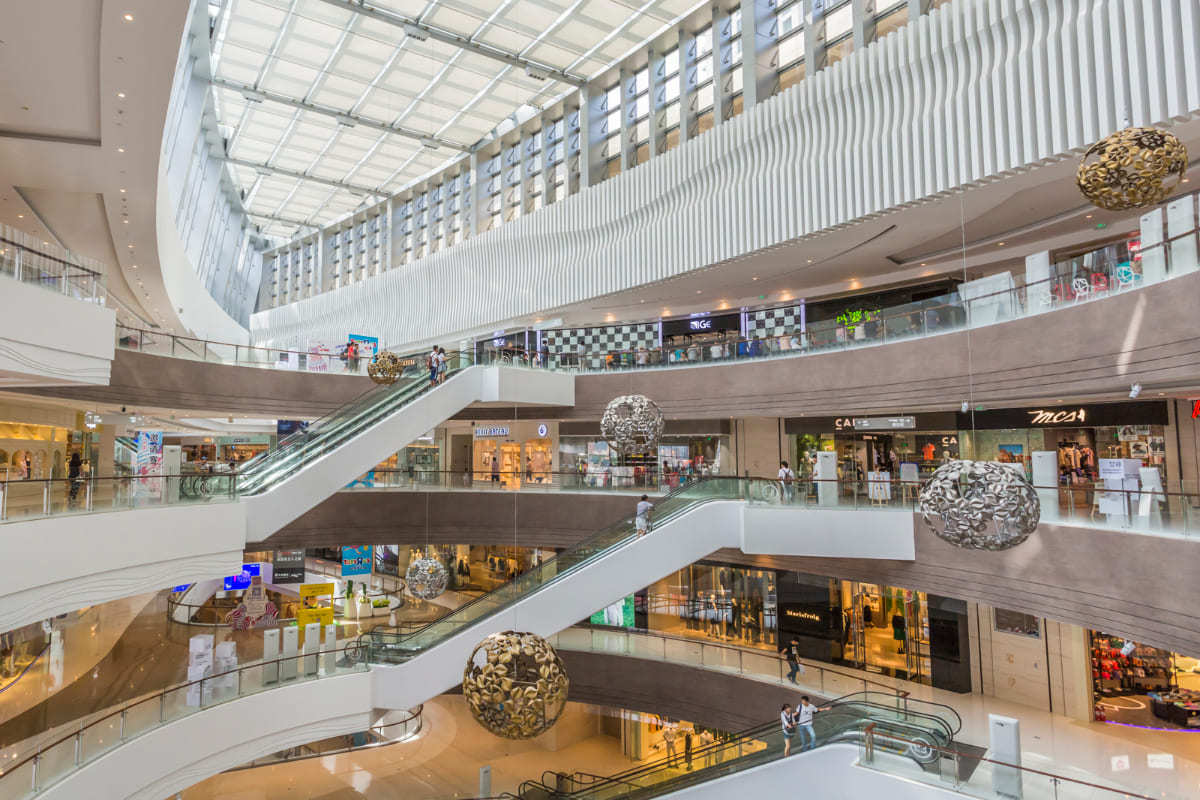 Interior de grande shopping center com pessoas transitando por seus andares e usando as escadas rolantes e várias luminárias artesanais penduradas como se caíssem do teto do prédio e ao fundo vários pontos comerciais com portas de vidro onde pode ser aberta uma Pop-up store