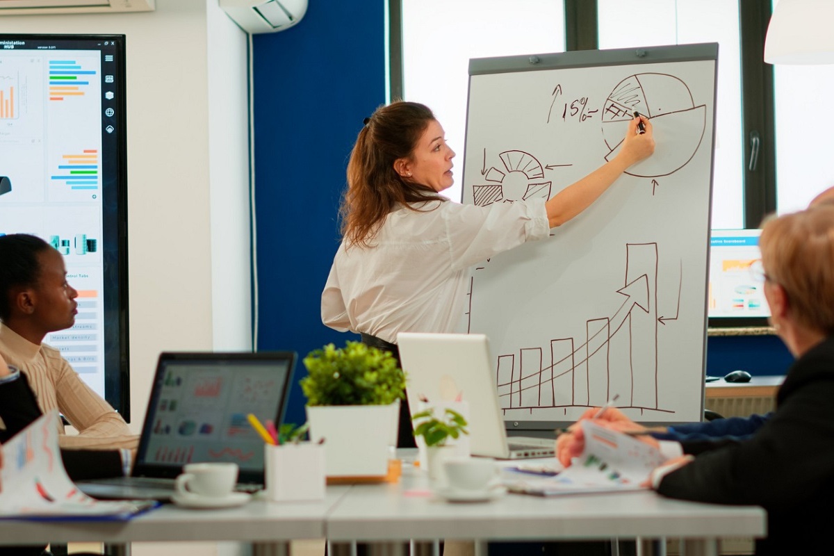 Mulher desenhando gráficos em painel branco durante reunião diante de colegas